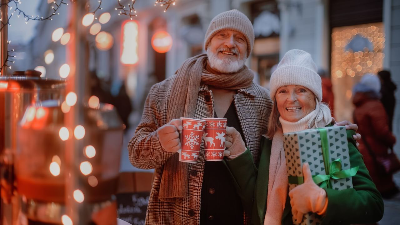 Elderly couple holding mugs and a gift, smiling, at a festive outdoor market.