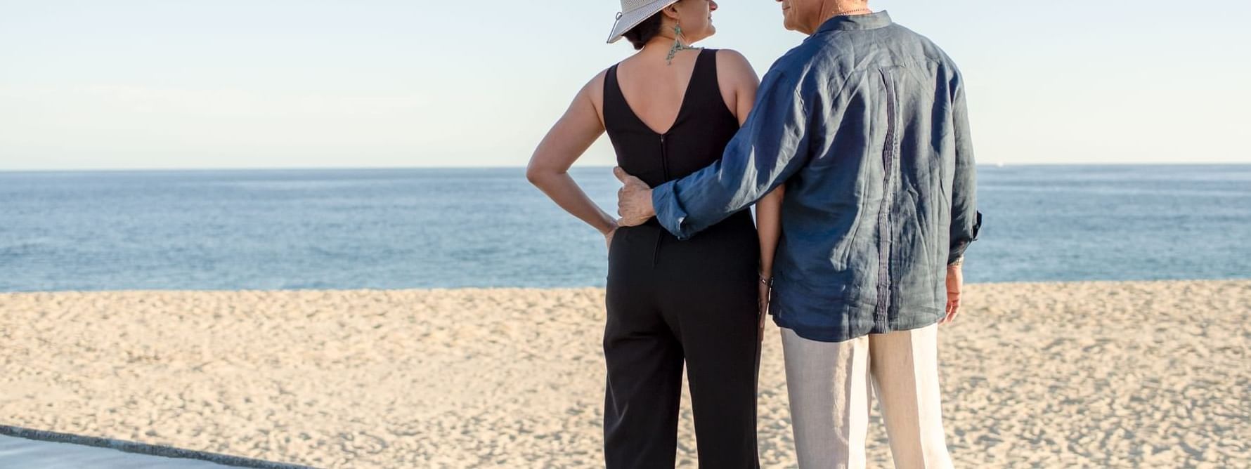 A couple in hats stands arm-in-arm facing the ocean on a beach, at the Marquis Los Cabos Resort