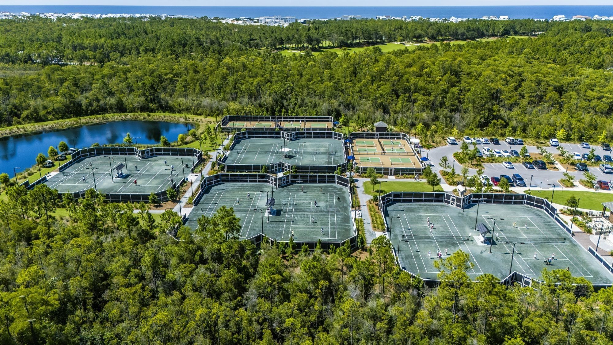 Aerial view of a tennis complex with multiple courts, surrounded by trees and a lake, near a parking lot with cars.