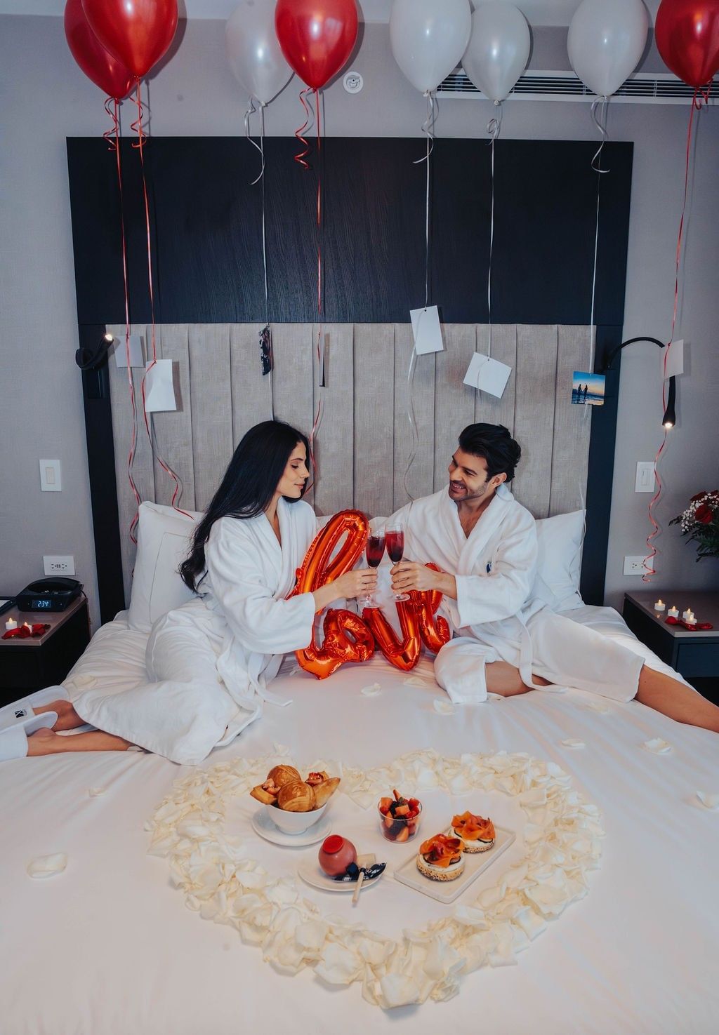 Couple toasting wine glasses by a love balloon, surrounded by petals and pastries at Hotel X Toronto