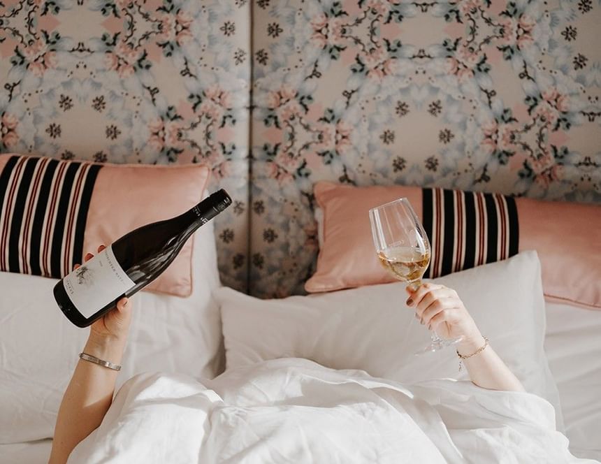 Woman in bed with white linens, holding a wine glass and wine bottle, floral headboard in background.