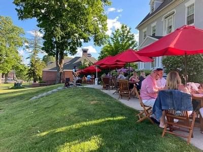 Guests dine at wooden tables under red umbrellas, by a live band near a white colonial building at Simsbury 1820 House