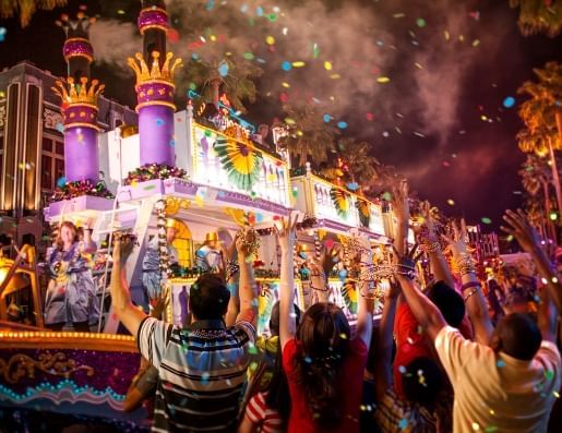 A large purple, green, and yellow float in a nighttime parade passing by a crowd of people with their hands in the air. 