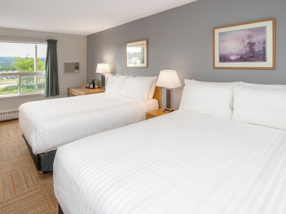 Two beds with white linens, lamps, a window, a table, and two framed paintings on a gray wall.