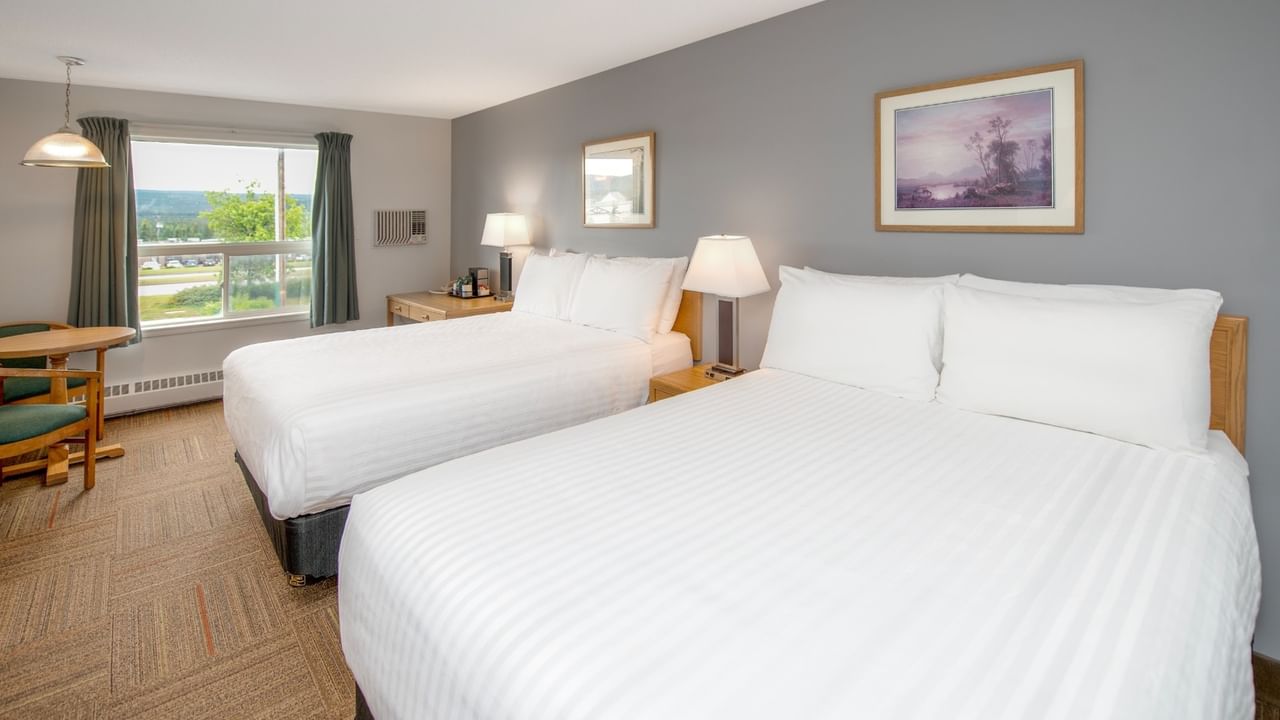Two beds with white linens, lamps, a window, a table, and two framed paintings on a gray wall.