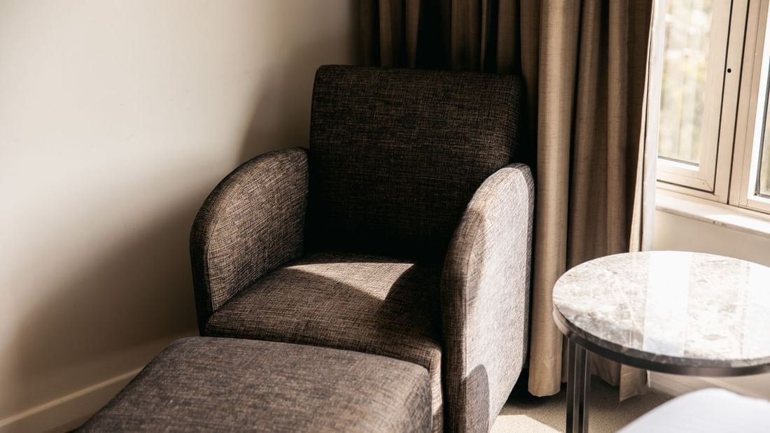 Armchair with footrest next to a marble side table by a window with curtains.