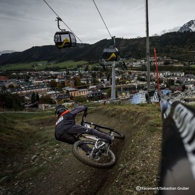 Mountainbiker fährt auf schlammigem Trail, mit Gondelbahn und Stadt im Hintergrund.