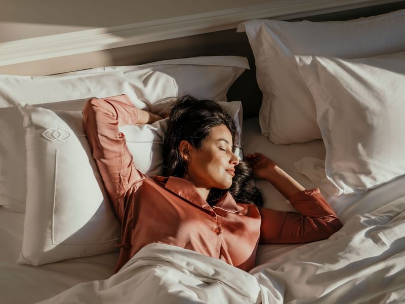 Woman in coral pajamas relaxing peacefully on white bedding in sunlit hotel room for Signature Escapes offer.