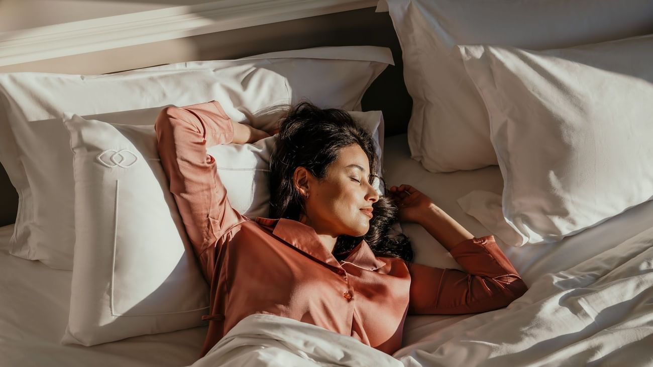 Woman in coral pajamas relaxing peacefully on white bedding in sunlit hotel room for Signature Escapes offer.