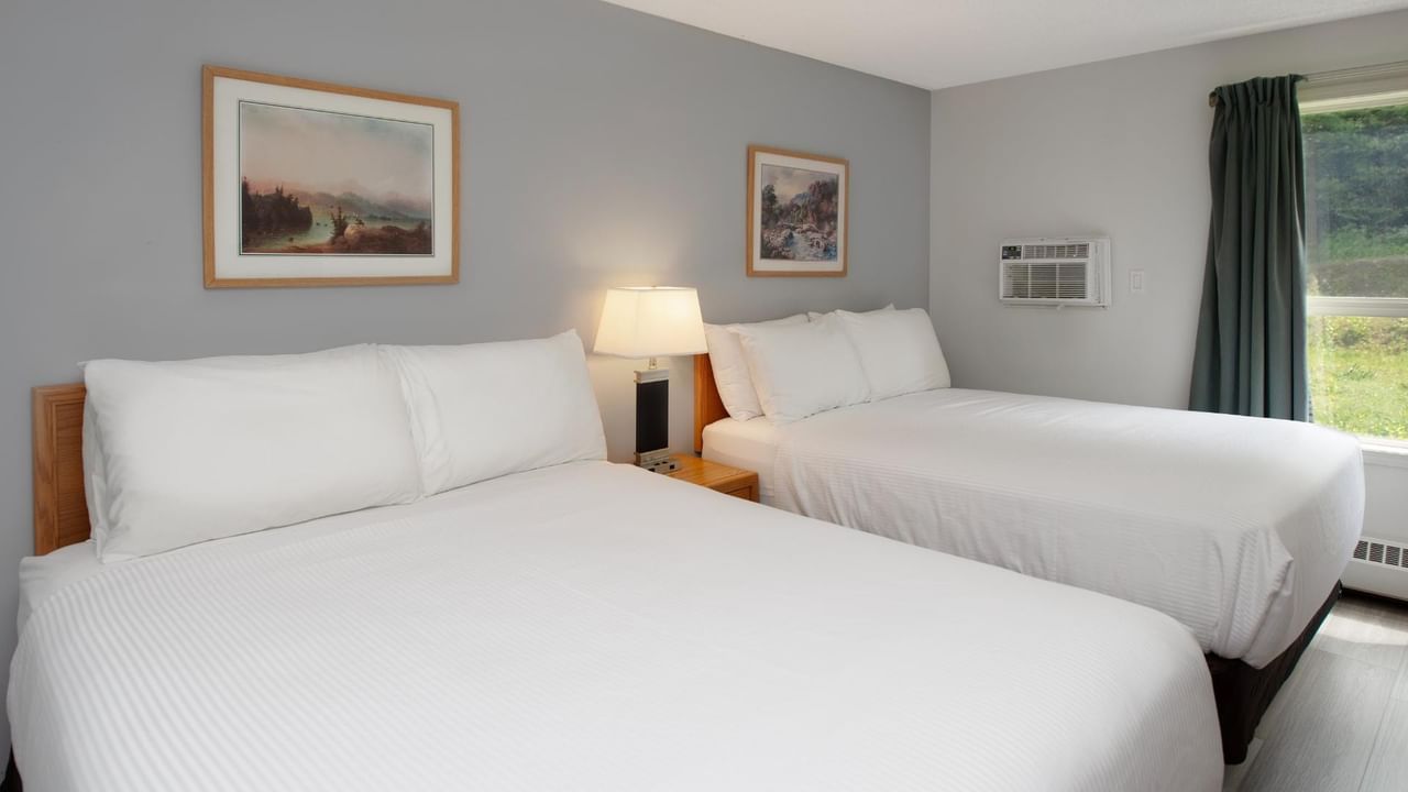 Two beds with white linens, pillows, nightstand with lamp, and framed art on gray walls.
