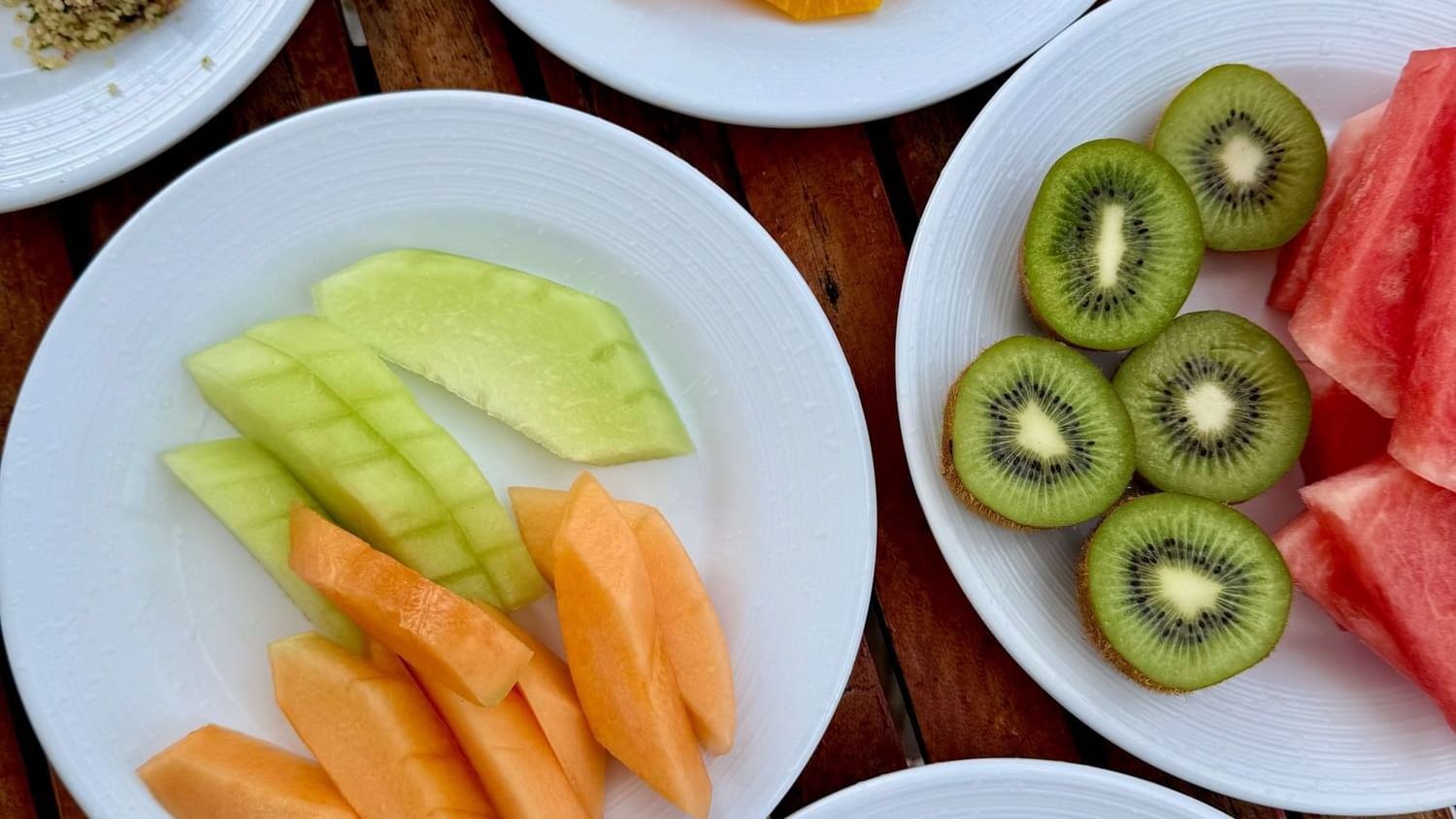 Five white plates with various fruits, an omelette, and garnishes on a wooden table.