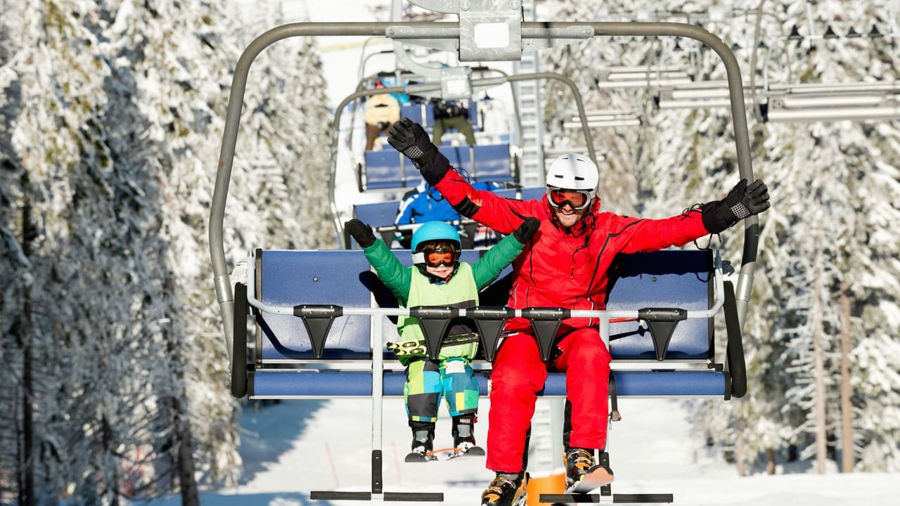 A father and child riding a ski lift, dressed in vibrant winter gear, with a snow-covered forest in the background.