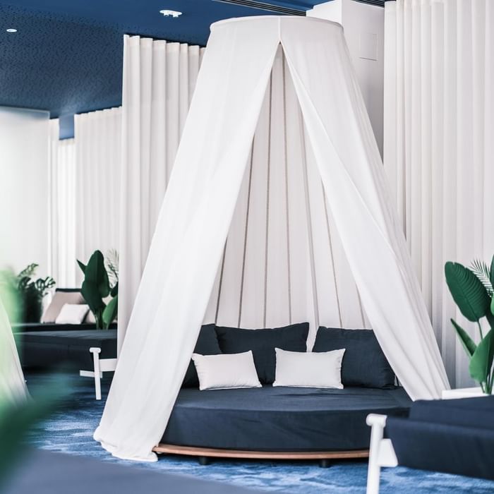 A circular bed with a white canopy, white pillows, and blue bedding in a room with white curtains and blue carpet.