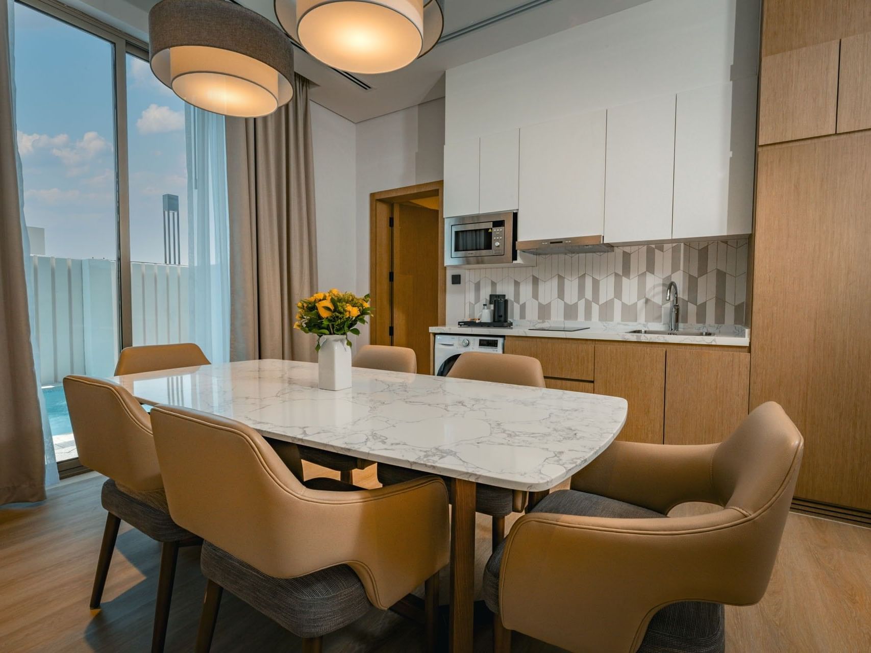 Dining room with beige chairs, marble table, and modern kitchen with white cabinets and wooden cabinets.