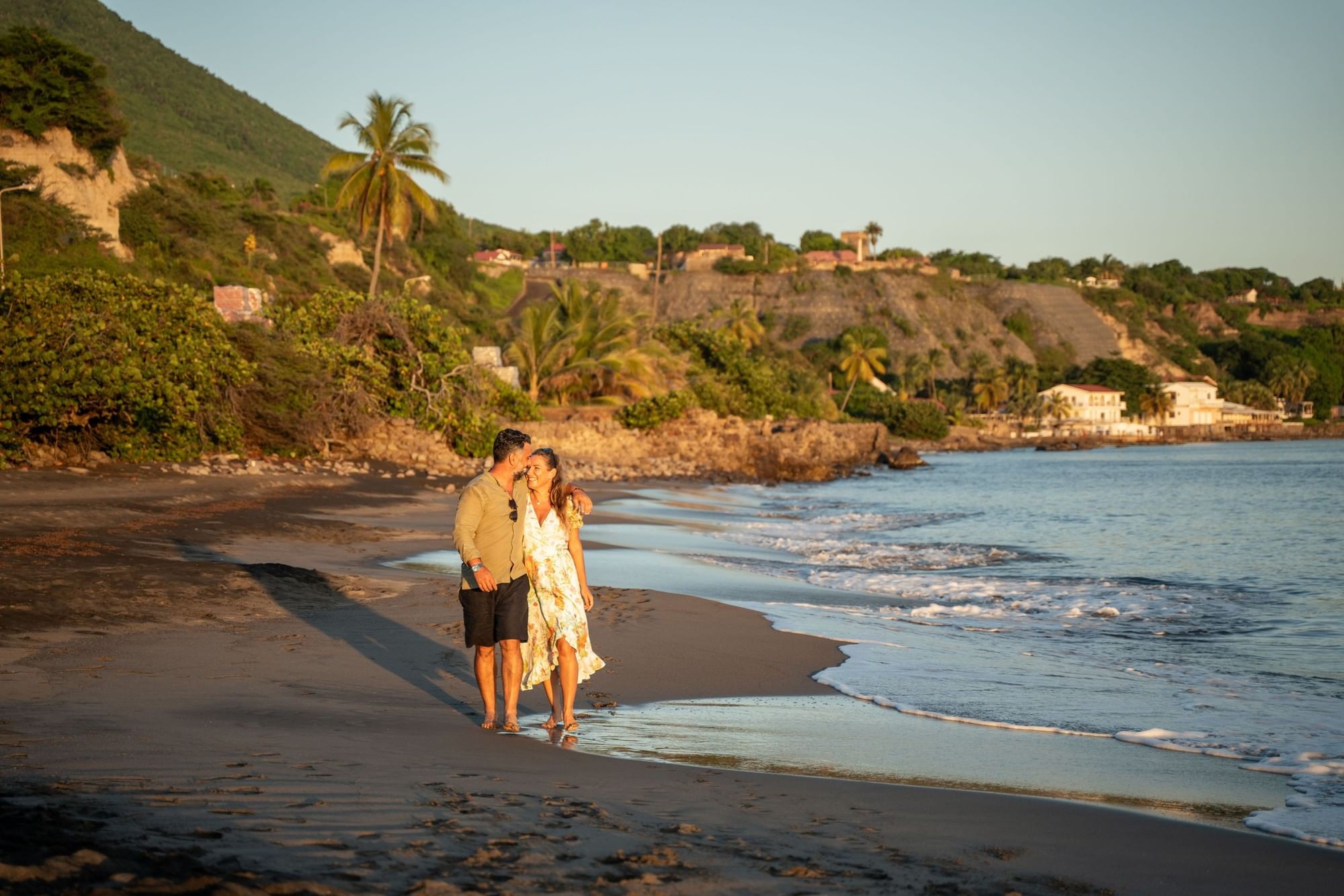 Couple walking together along a peaceful dark sandy shoreline near gentle waves at sunset at Golden Rock Resort
