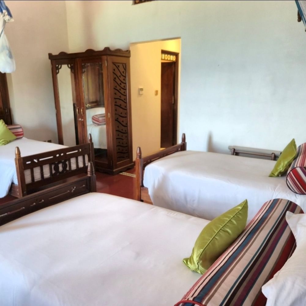 Three wooden beds with white linens and colorful pillows in a Standard Triple Room at Zanzibar Serena Hotel.