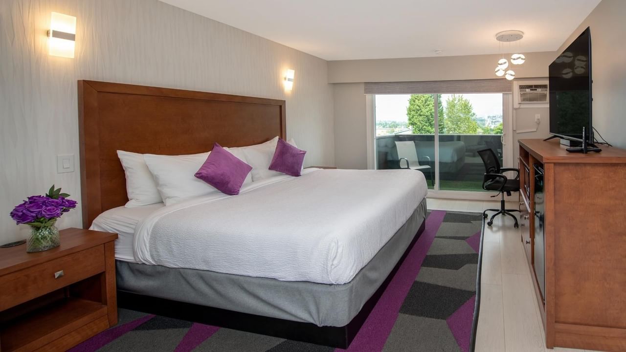 A bedroom with a king bed, nightstand, purple pillows, TV, desk, chair and balcony.