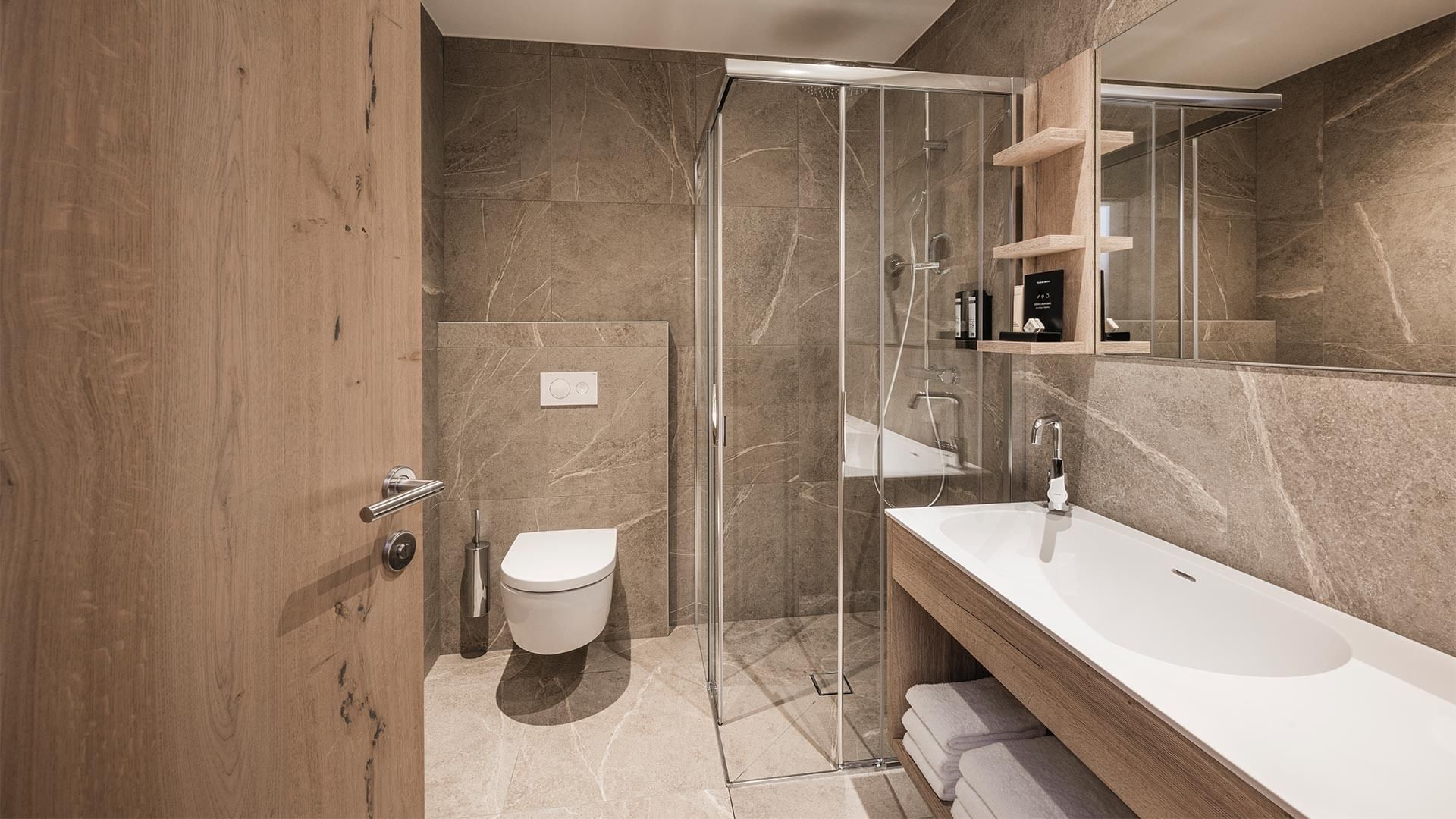 Badezimmer mit Holztüren, marmorierter Wand, Glasdusche, WC und Waschbecken mit Spiegel.