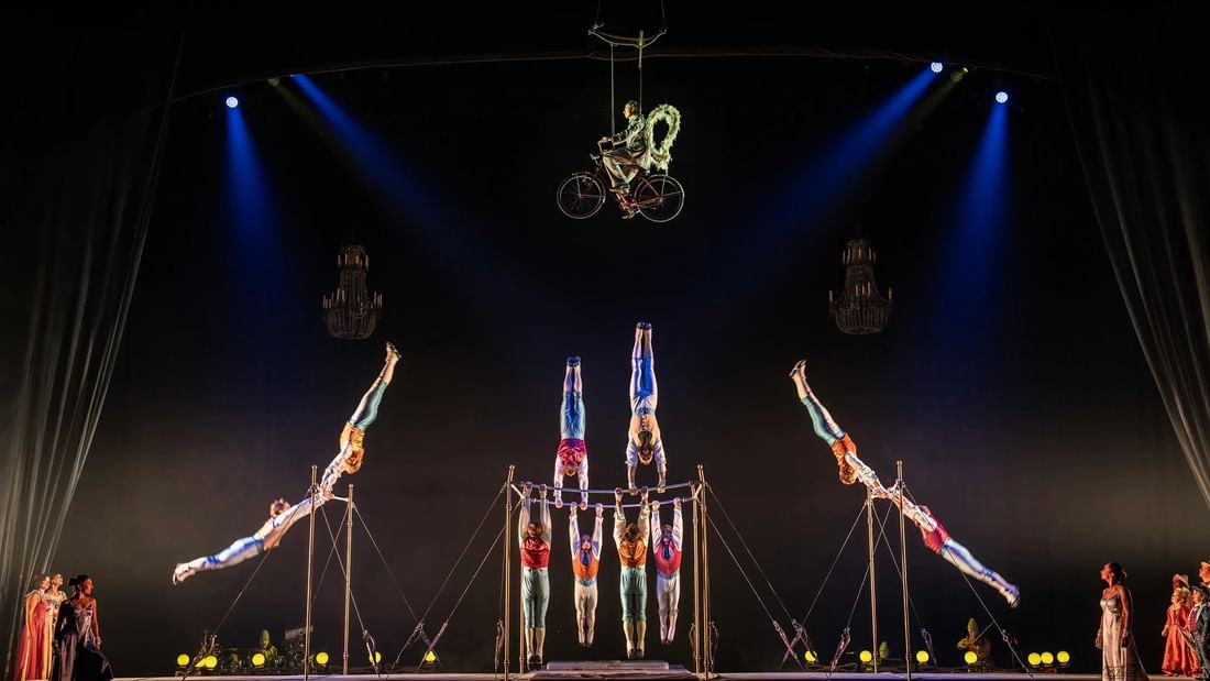 Acrobats perform on stage with trapezes, one cyclist riding a bike mid-air, under blue spotlights.