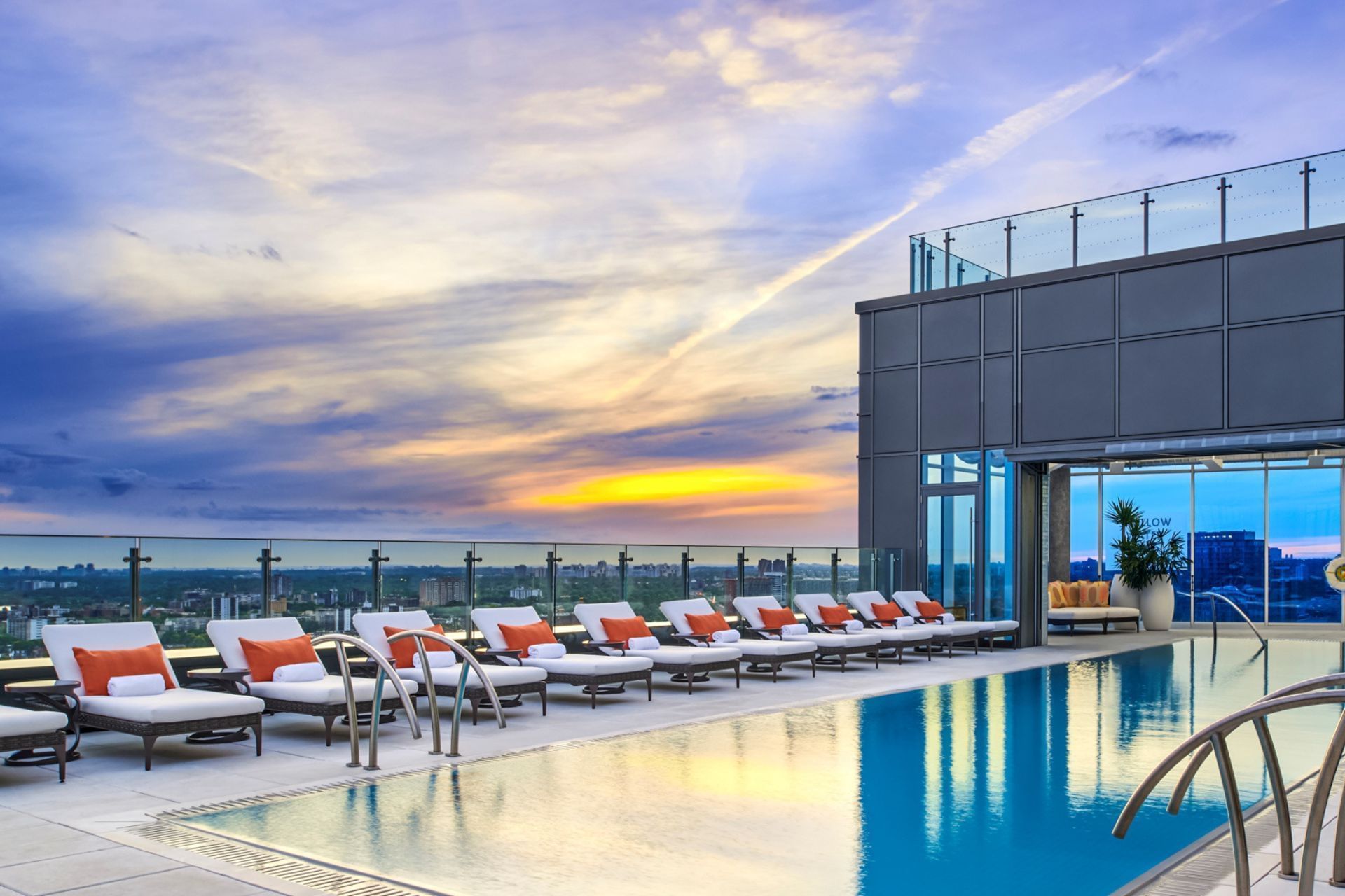 Rooftop pool with lounge chairs, large windows, and view of sunset over city skyline.