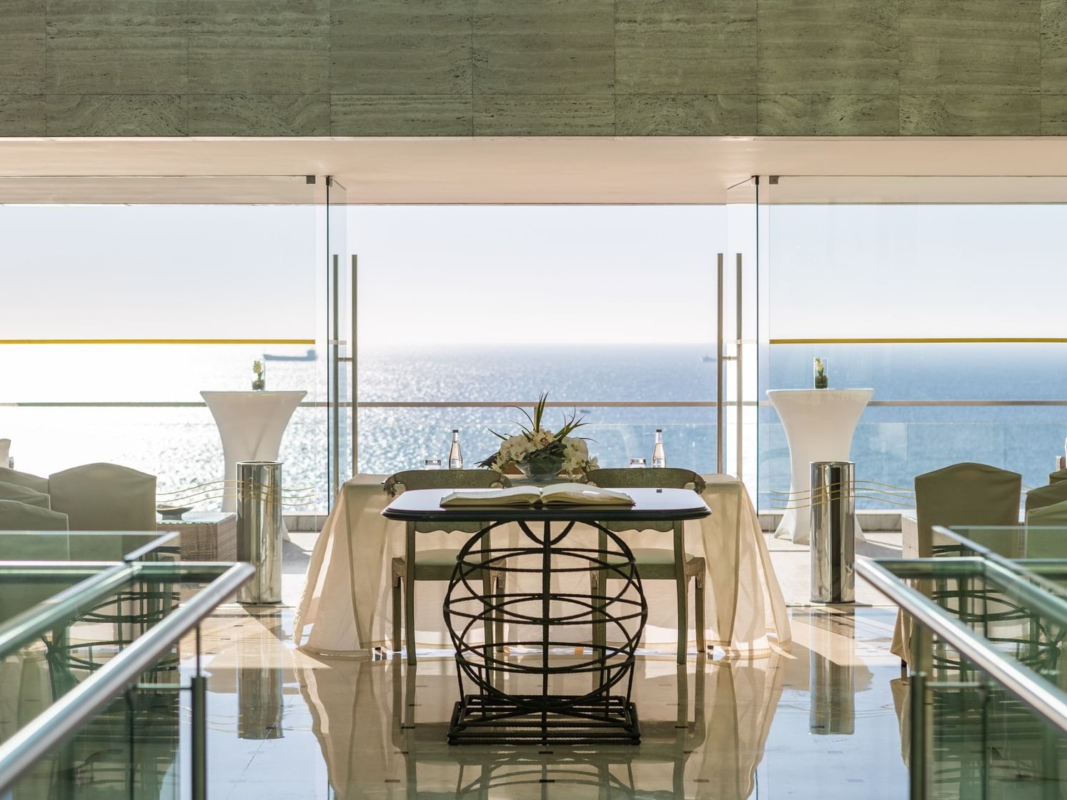 Sala de bodas con vista al mar, mesa central con libro y flores, sillas y barandas de vidrio.