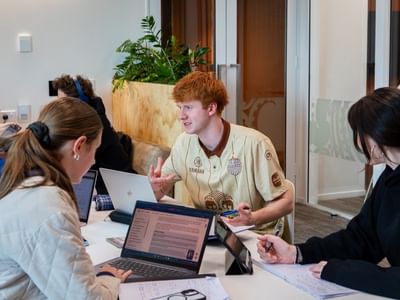 Hotel staff member in beige uniform assisting guests at modern reception desk with laptops and wooden planter nearby.