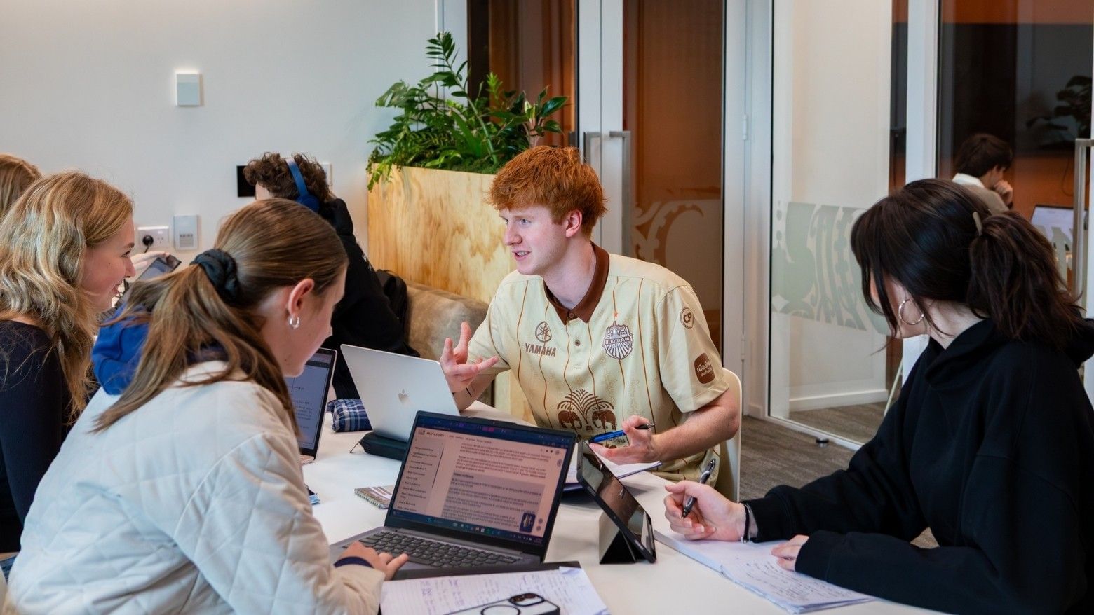 Hotel staff member in beige uniform assisting guests at modern reception desk with laptops and wooden planter nearby.