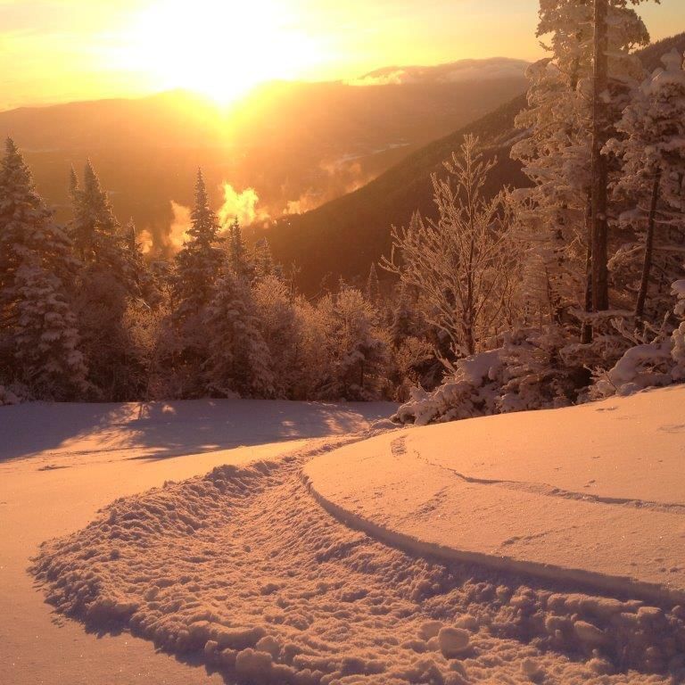Snowy mountain landscape with sun setting behind hills and evergreen trees in winter.