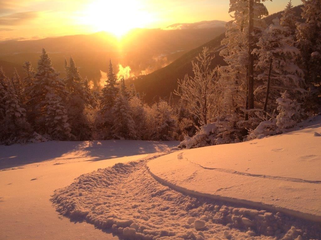 Snowy mountain landscape with sun setting behind hills and evergreen trees in winter.