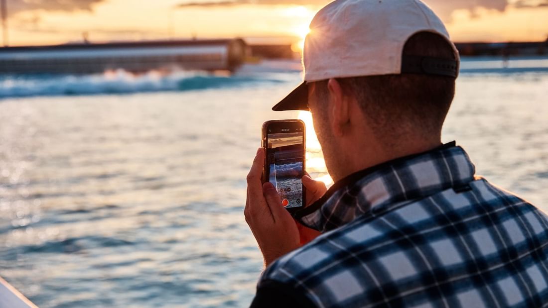 Man capturing a seaside sunset on their smartphone at URBNSURF Sydney near Novotel Sydney Olympic Park
