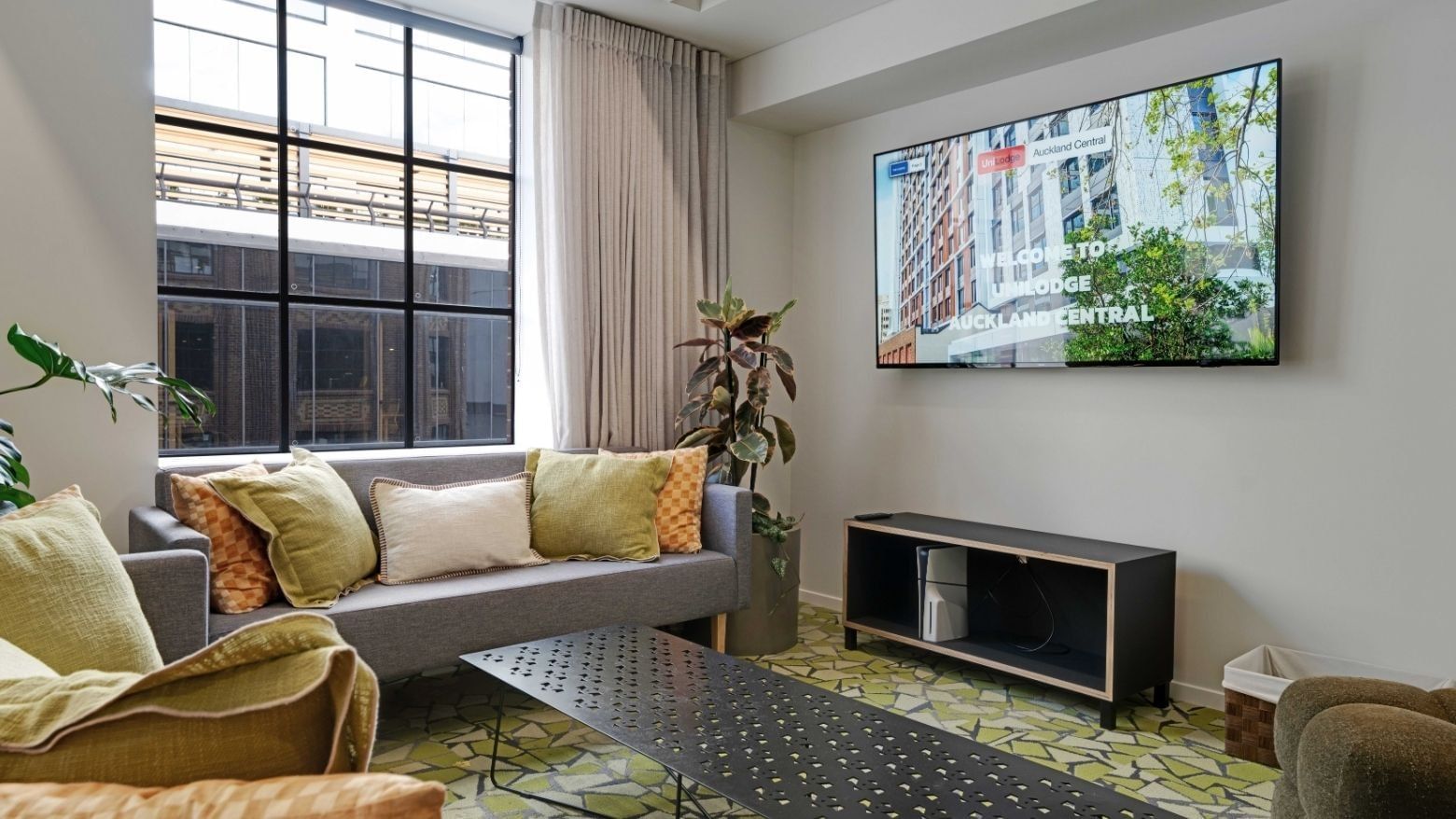 Modern living room with gray sofa, yellow cushions, large windows, and wall-mounted TV at UniLodge Auckland Central