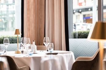 Elegant dining table setting with wine glasses, a brass lamp, and soft blush curtains by a window at Téséo Restaurant & Bar