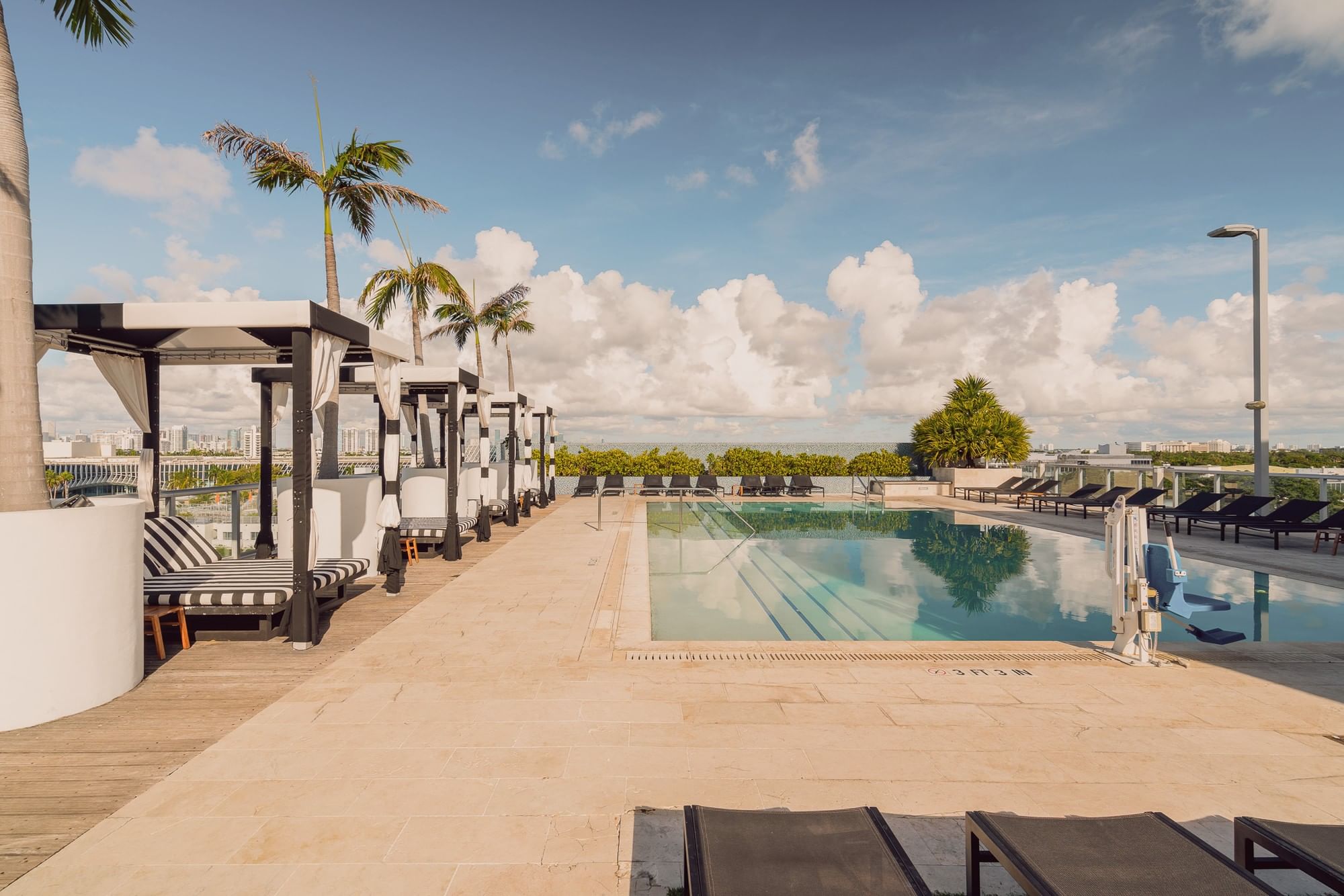 A rooftop pool area with a rectangular pool, lounge chairs, cabanas, and palm trees overlooking the city.
