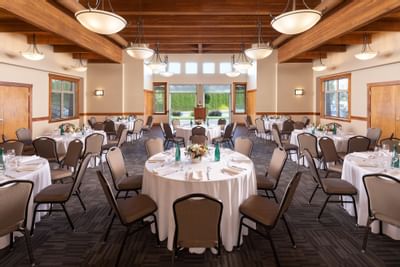 A large dining hall with multiple round tables set with white cloths, chairs, and floral centerpieces under wooden beams.