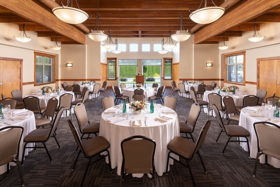 A large dining hall with multiple round tables set with white cloths, chairs, and floral centerpieces under wooden beams.