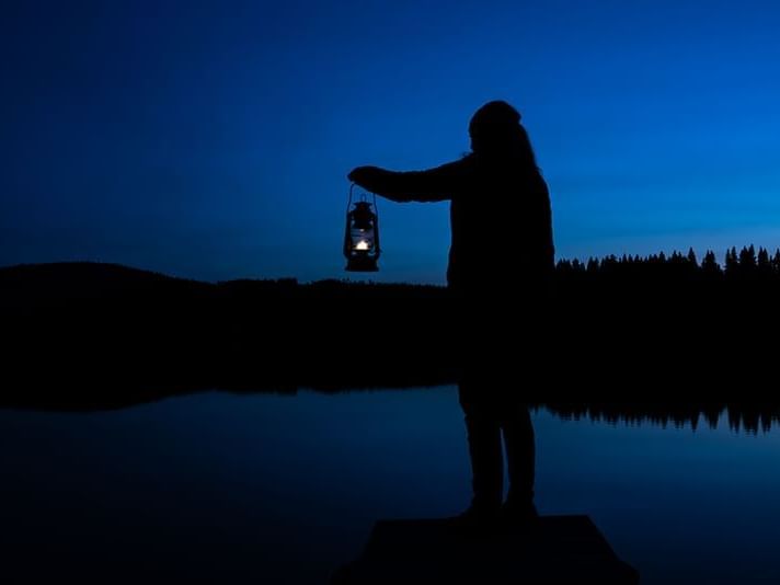 Silhouette of a person looking into night by a lake, holding a lit lamp.