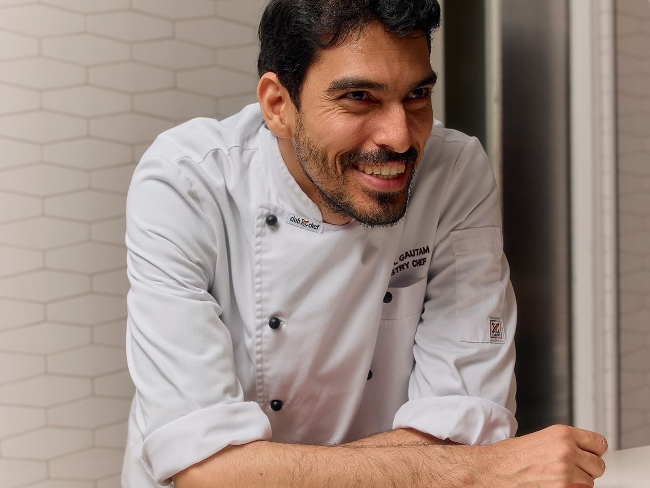 Chef Gautam in white uniform smiling behind counter with two plates of food.