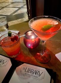 Two colorful cocktails garnished with lime and berries beside The Library Bar\'s speciality cocktails menu