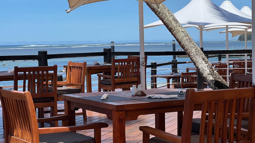 Outdoor dining area with wooden tables and chairs under umbrellas overlooking the ocean at Warwick Fiji Resort and Spa, Korolevu.