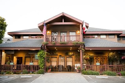 Front view of a lodge with flags, flowers, patio, and chairs, surrounded by greenery and a parking area.