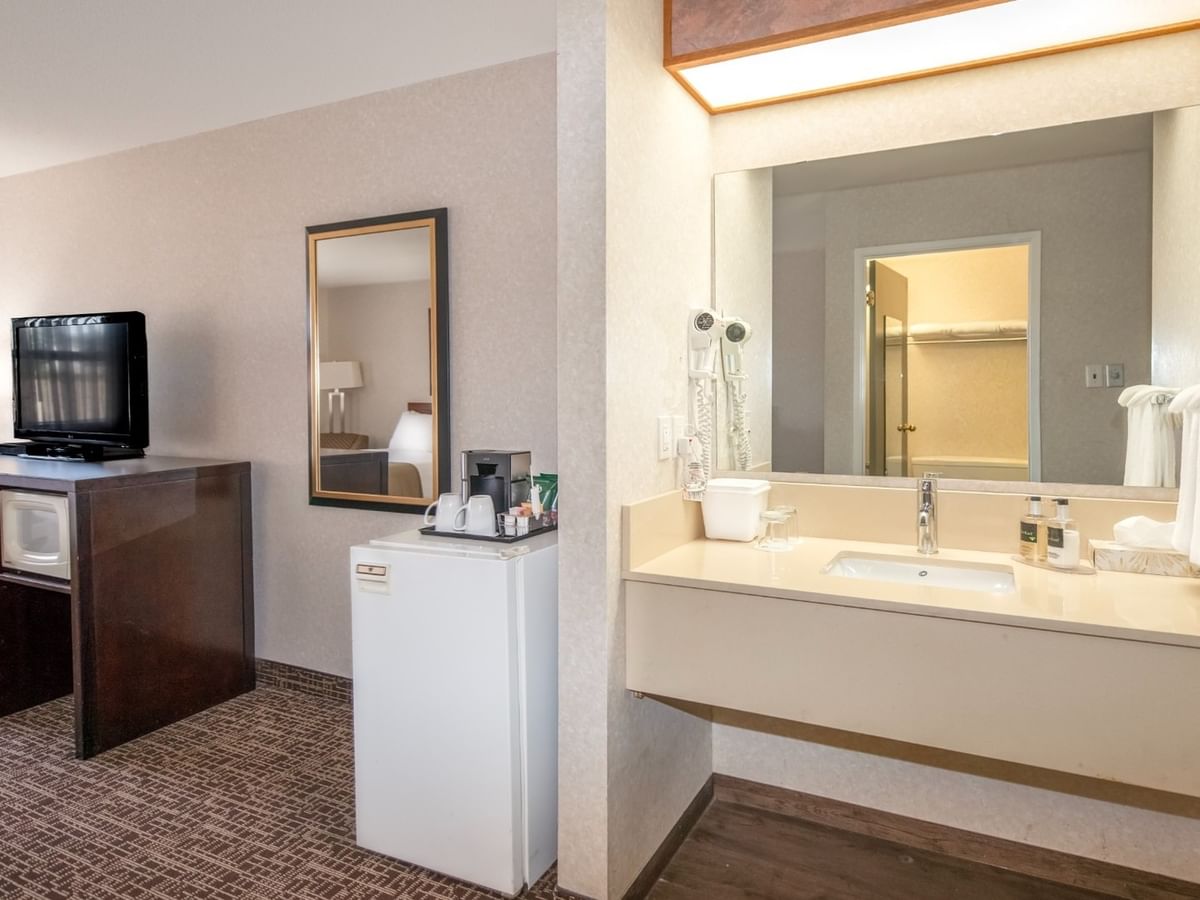 Hotel room with TV, dresser, mirror, mini-fridge, and bathroom with sink and mirror.