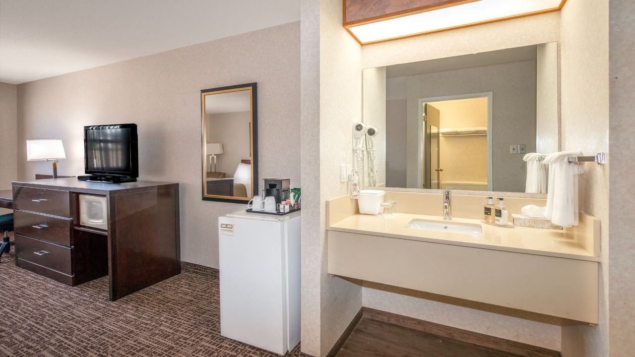 Hotel room with TV, dresser, mirror, mini-fridge, and bathroom with sink and mirror.