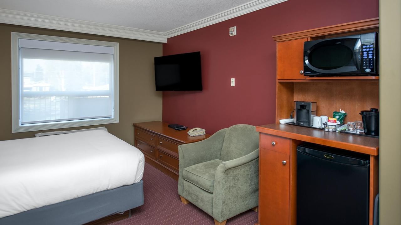 Hotel room with white bed, sage green armchair, kitchenette with microwave and mini-fridge at Coast Edson Hotel.