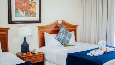 Standard Room Double featuring two beds with blue linens, a floral artwork, & a bedside table at Bay Gardens Inn