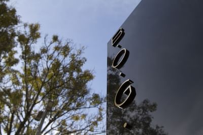 Cartel vertical negro con texto y símbolos en relieve, junto a un árbol y cielo azul.