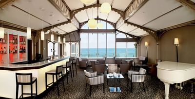 Bright bar area featuring white piano and cozy seating with stunning ocean views at Le Grand Hôtel des Thermes