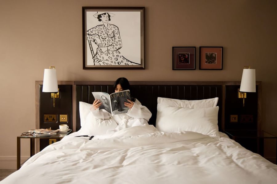 Woman relaxing on a cozy bed while reading a fashion magazine in the Collection Superior Room at The May Fair Hotel