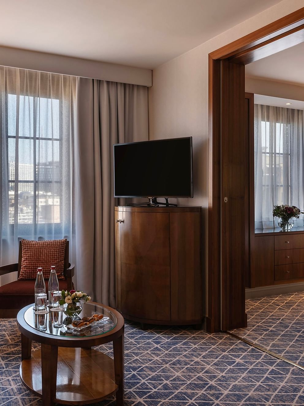 Spacious hotel suite with separate living area, wooden furnishings, large windows, and modern amenities in warm tones.