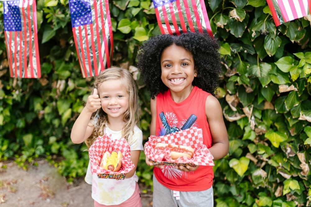 A boy and a girl in patriotic shirts smile holding hot dogs in front of a green shrub decorated with American flags.