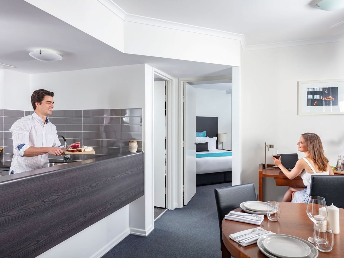 Spacious hotel apartment with a man cooking in the kitchen and a woman working at a desk at The Sebel Brisbane